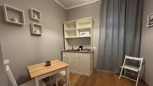 A compact kitchen with white cabinets, a wooden table, and a small bench. There is a white chair and a shelf unit with a coffee maker. - Gravina Suite Frattina (Rome)