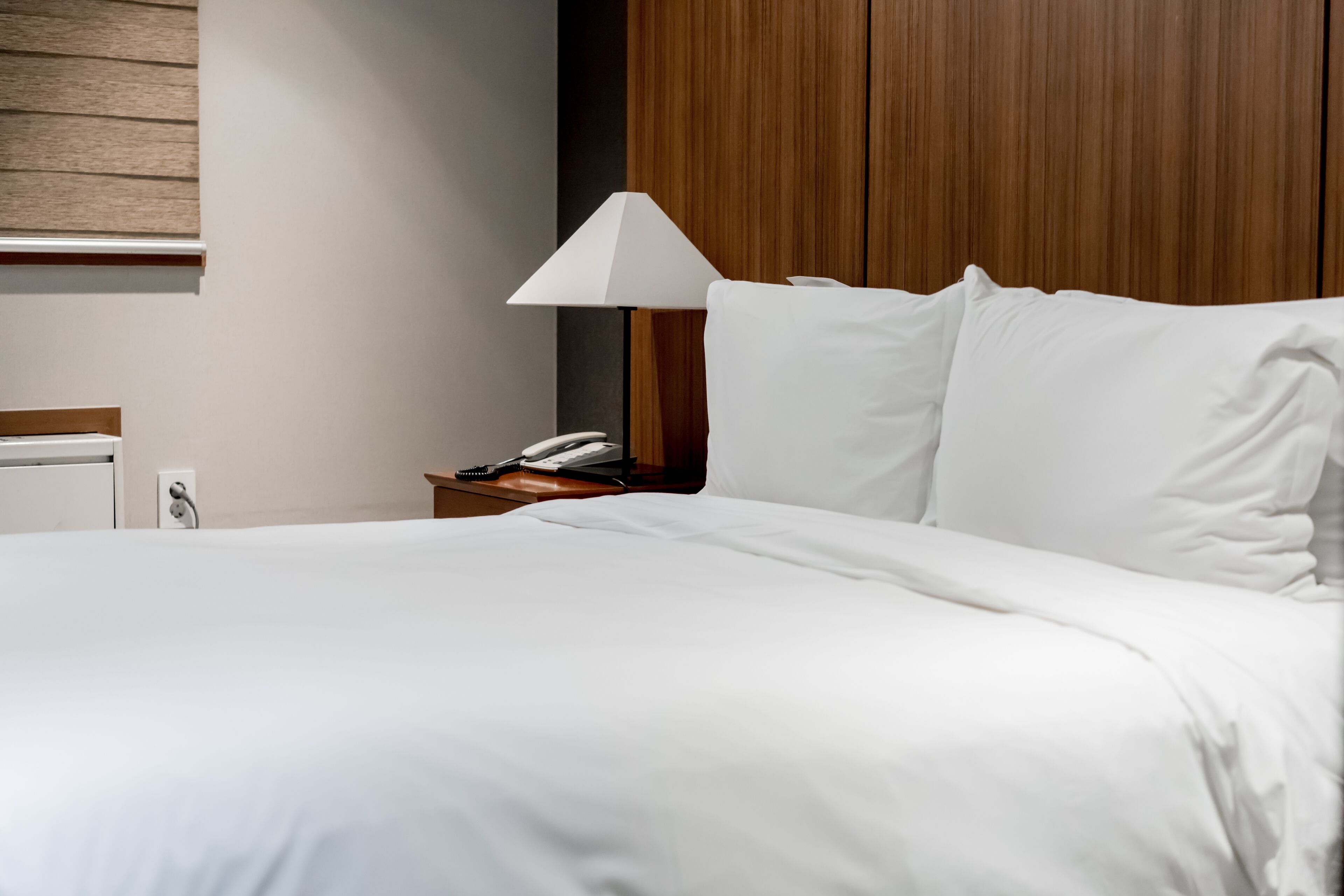 Una cama bien tendida con sábanas blancas, una mesita de noche con lámpara y un cabecero de madera.