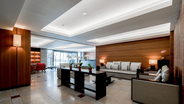 Un moderno vestíbulo de hotel con un sofá, una mesa de centro y sillas. Se observa una pared decorativa de madera y un gran ventanal que proporciona luz natural.