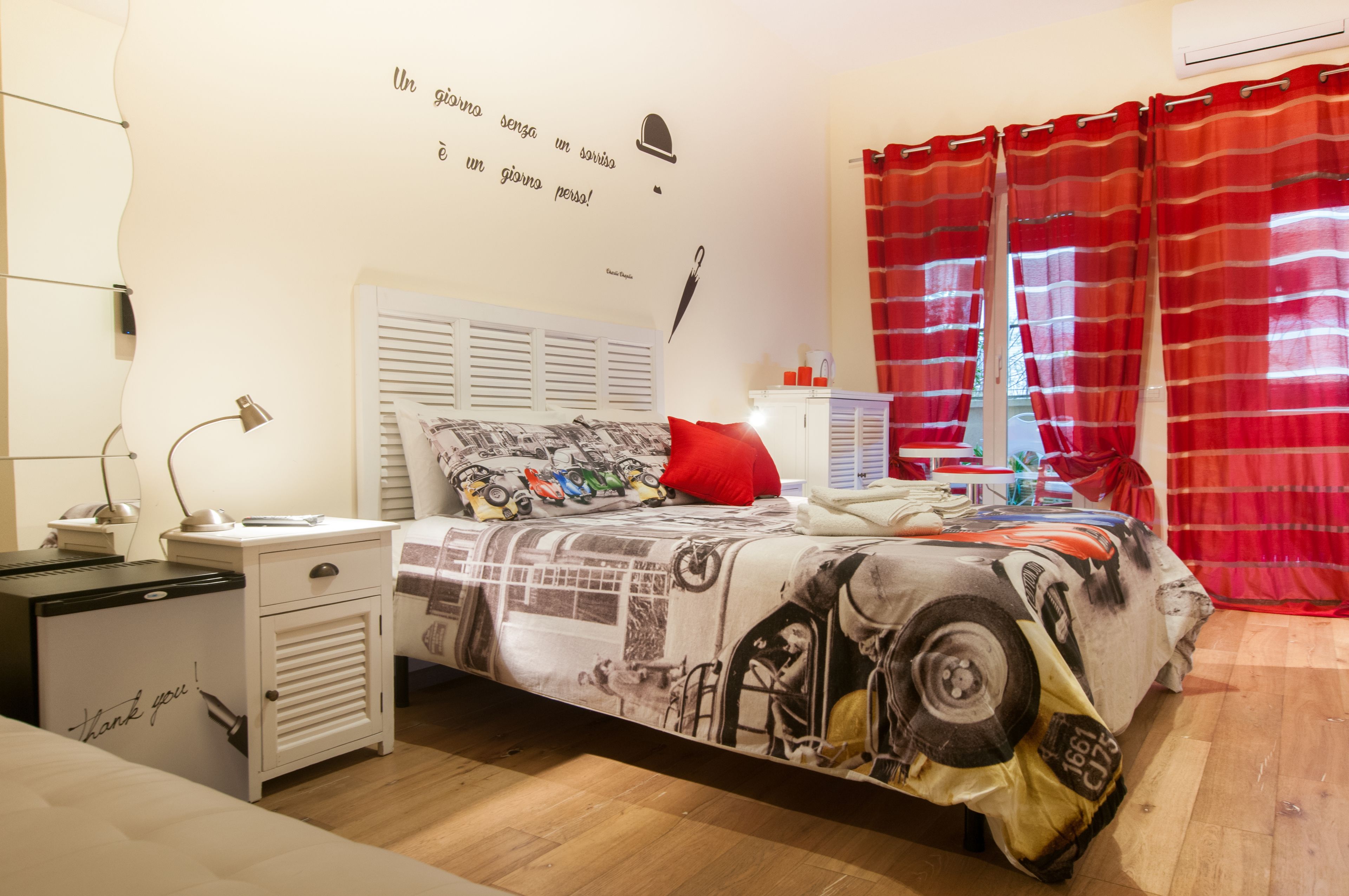A bedroom with a bed featuring a cityscape print, a white bedside table with a lamp, a red pillow, and a wall with a quote.
