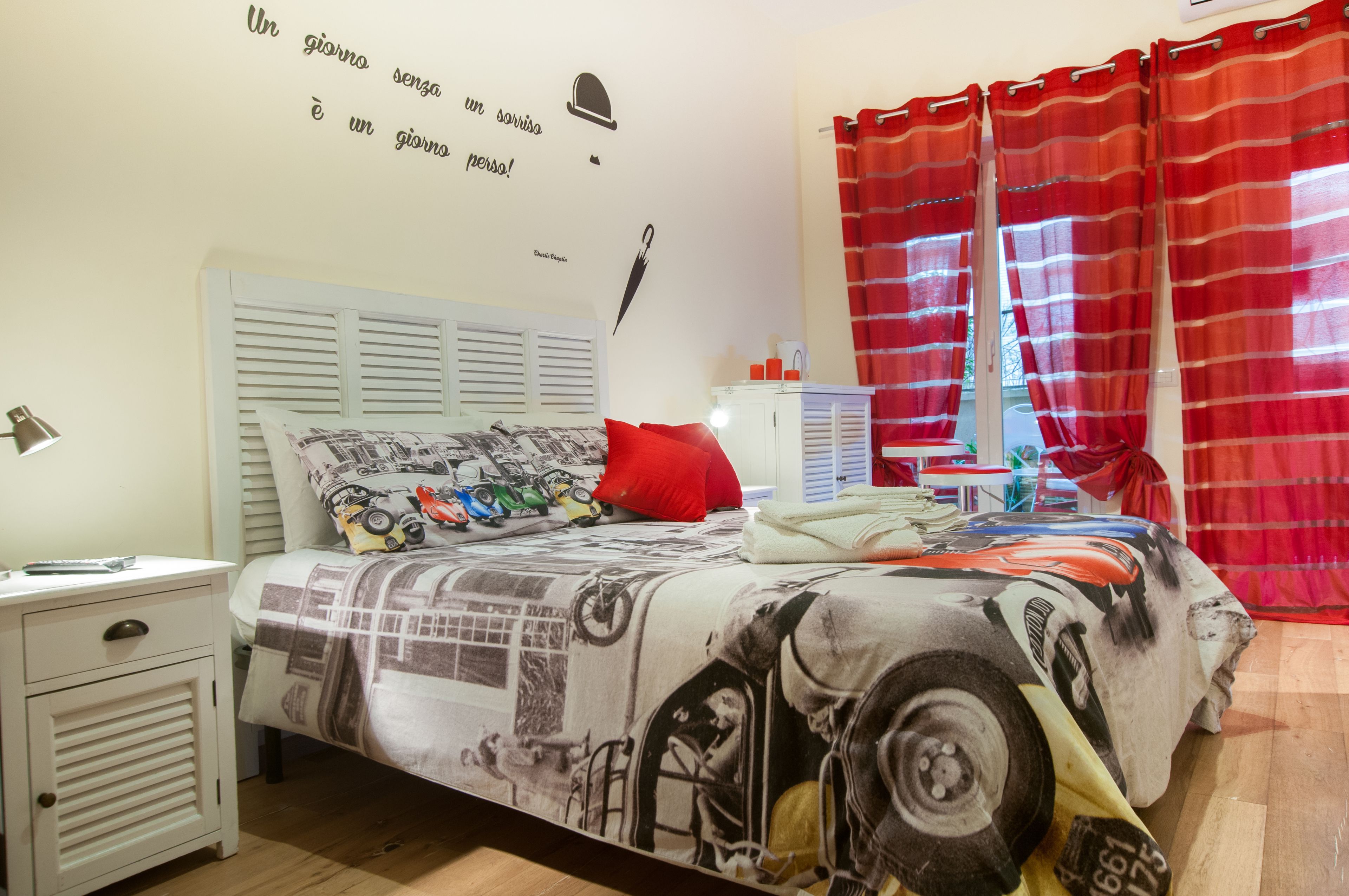 Un dormitorio con cama, mesita de noche, lámpara, ventana con cortinas rojas y pared con citas motivacionales.