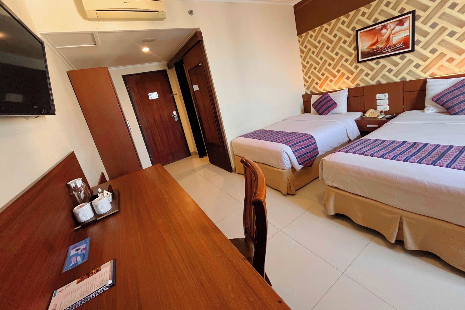 A hotel room with two beds, a desk with a chair, a TV, and a patterned wall.