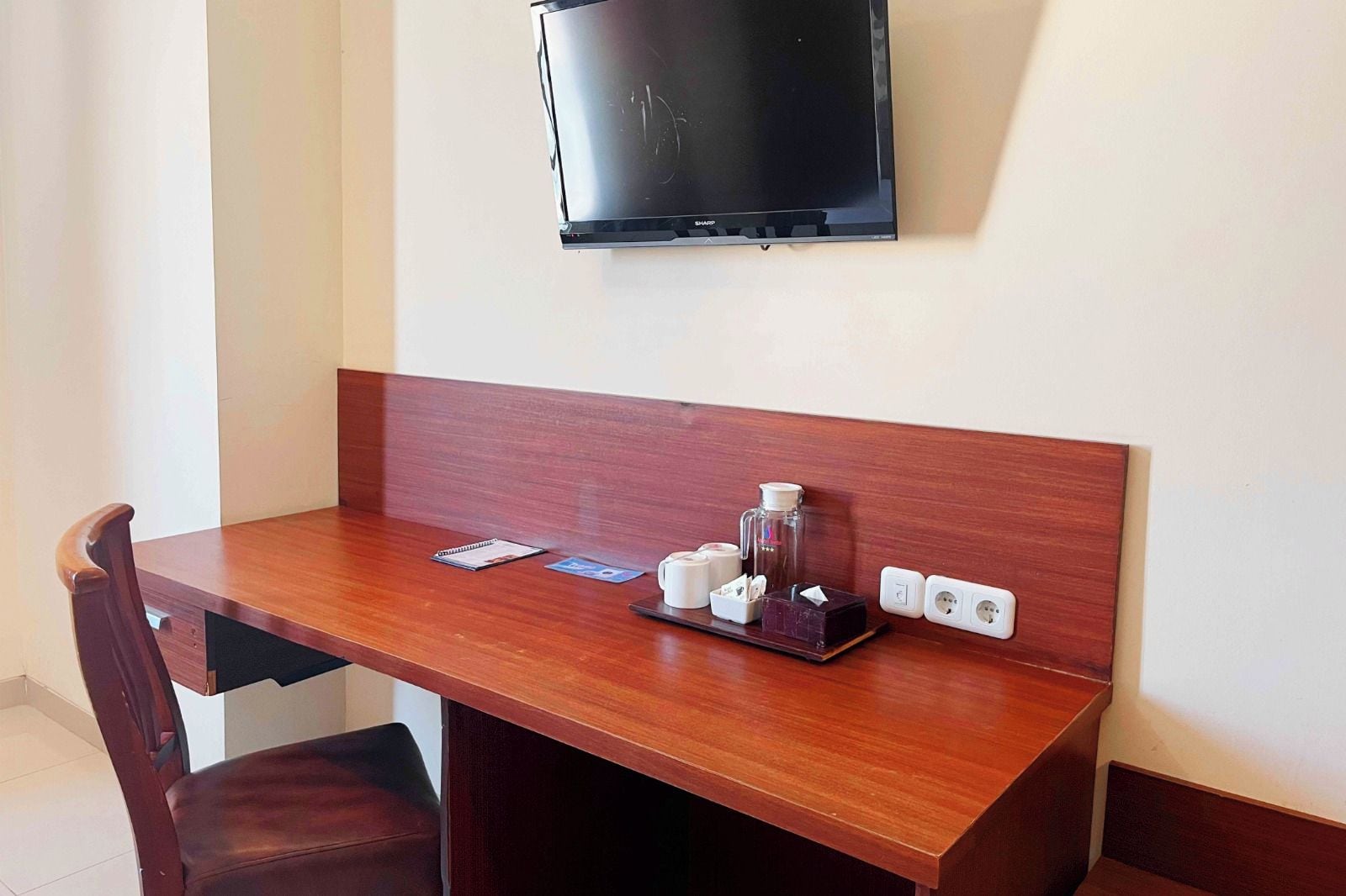 A wooden desk with a television mounted above it, a chair, and a tray with cups and a tissue box.