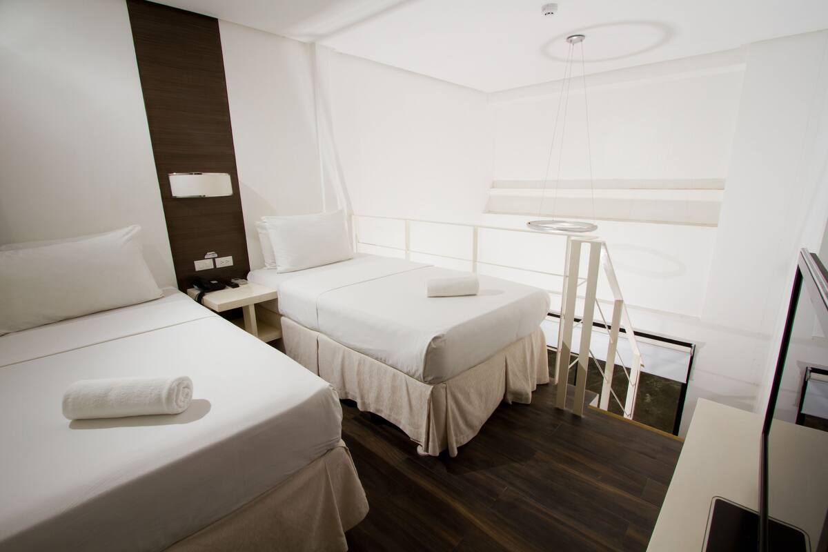 A hotel room with two beds, a small table, a chair, a desk, and a window.