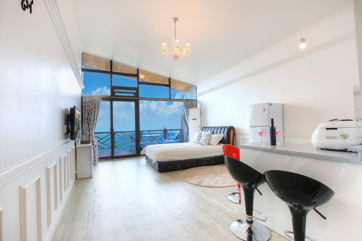 A modern hotel room with a large balcony, a bed, a dining area, and a kitchenette.