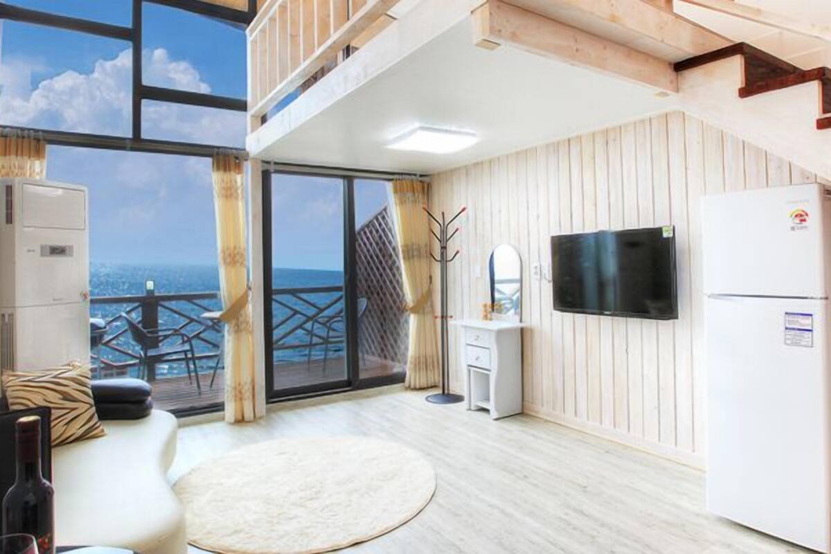 A modern kitchen with a large window overlooking the sea, a flat-screen TV, and a refrigerator.