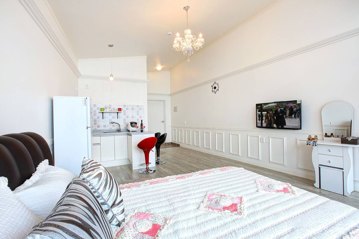 A modern hotel room with a large bed, a sitting area with a red chair, a kitchen area, and a bathroom with a mirror.