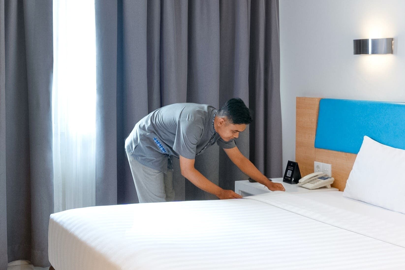 A hotel room with a neatly made bed, a bedside phone, and a wall-mounted light fixture.
