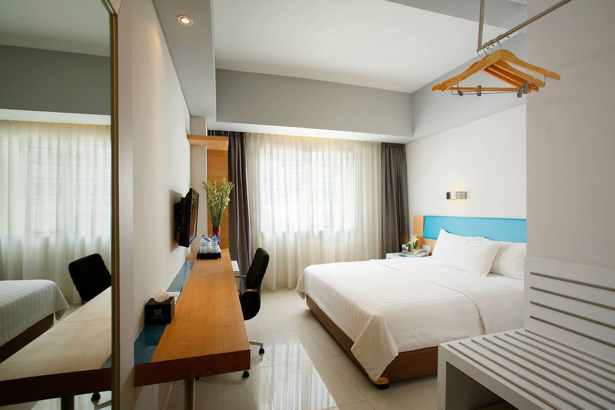 A hotel room with a bed, a desk, a chair, a TV, and a bedside table.