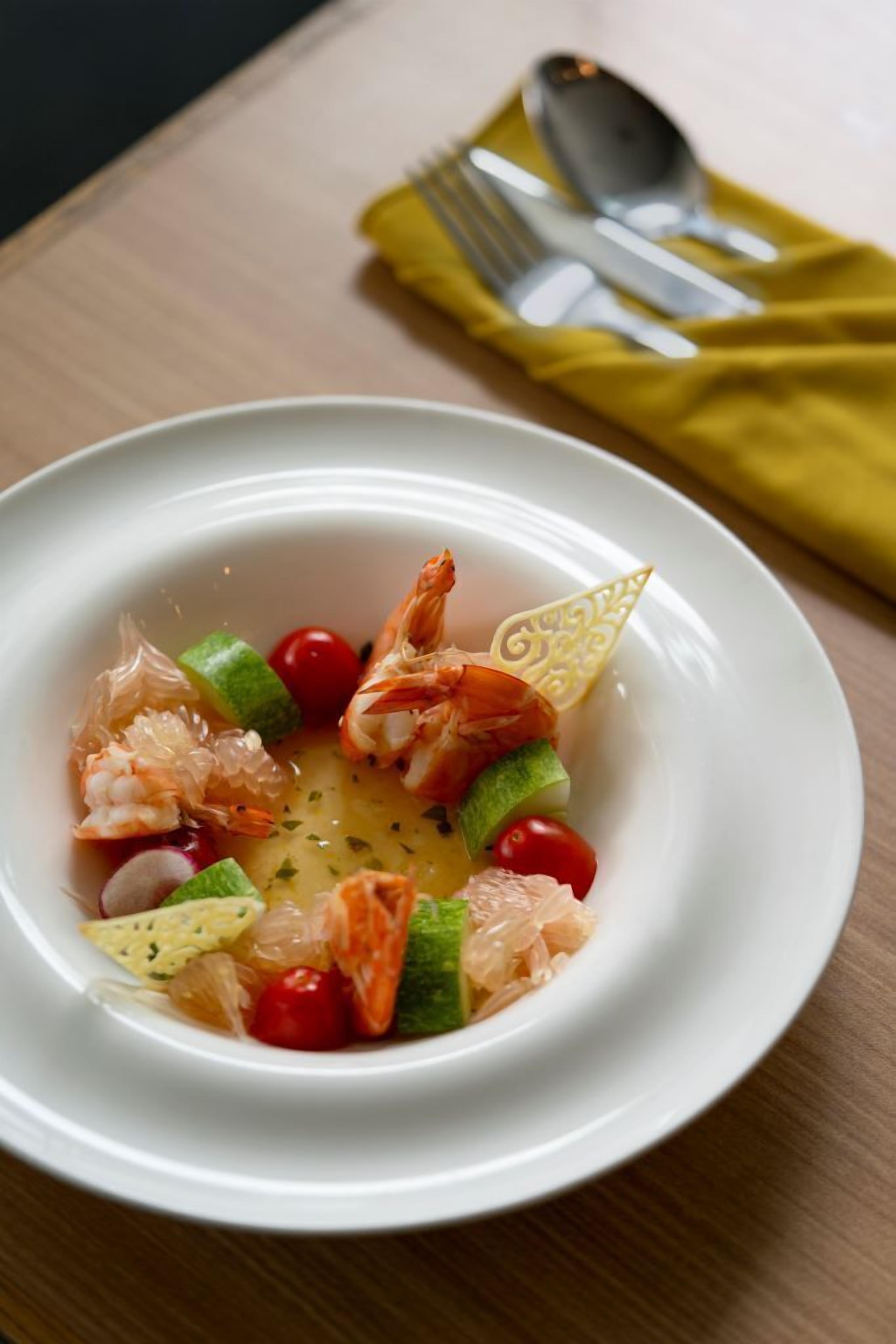 A white bowl of seafood salad with shrimp, cherry tomatoes, and cucumber slices on a wooden table.