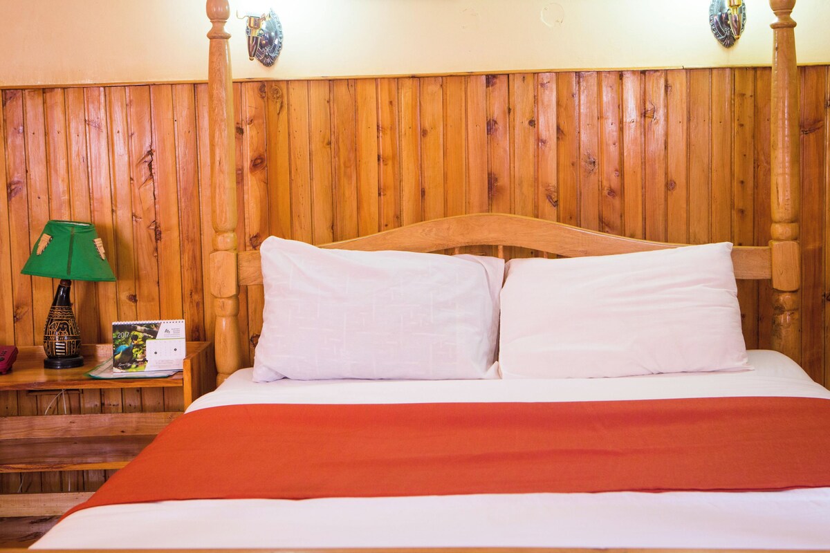A wooden-paneled room with a bed, a nightstand, a green lamp, and a decorative wall piece.