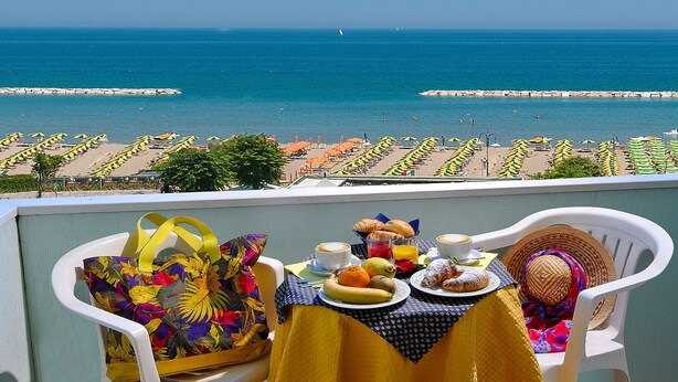Ein Balkon mit Frühstückstisch mit Blick auf einen Strand mit Sonnenschirmen und einem Felssteg.