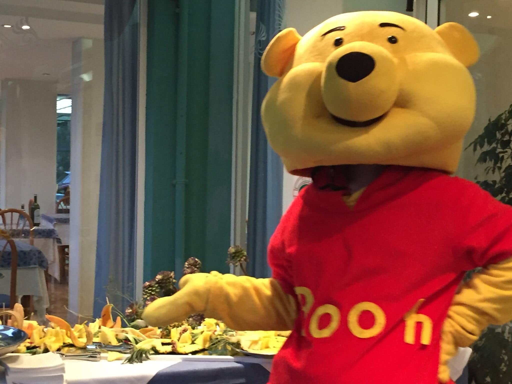 Une personne vêtue d’un costume d’ours jaune et portant un t-shirt rouge avec l’inscription « Winnie l’ourson », se tenant devant une table ornée d’un arrangement floral.