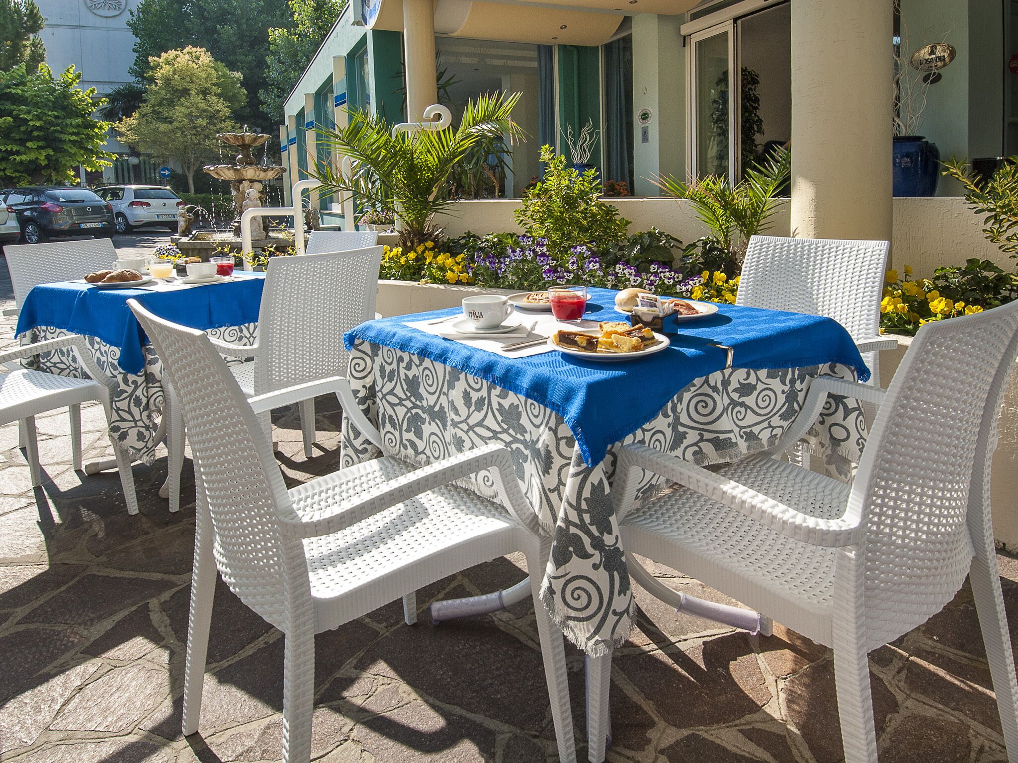 Un área de comedor al aire libre con sillas y mesas de mimbre blanco cubiertas con manteles azules, con una fuente y plantas en macetas al fondo.