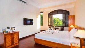 A hotel room with a bed, a TV, a desk, and a window with a view of greenery.