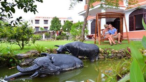 A garden with two large elephant sculptures in a pond, surrounded by greenery and a building with a red roof in the background.