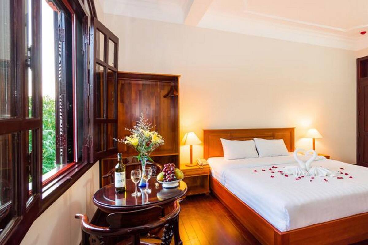 A hotel room with a large bed, wooden headboard, two bedside lamps, a small table with a vase of flowers, and a window with a view of greenery.
