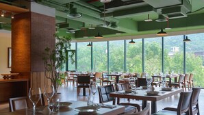 En restaurant med flere spiseborde og stole, store vinduer med udsigt over grønne områder, og et moderne industrielt loftsdesign.