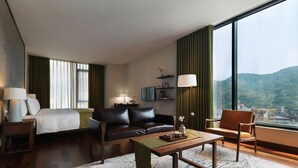 Et moderne hotelværelse med en seng, en lædersofa, et træbord og udsigt til et bjergrigt landskab gennem store vinduer.