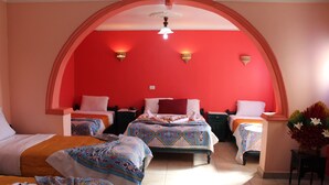 Ein Hotelzimmer mit drei Betten, jedes mit einer gemusterten Tagesdecke und weißen Kissen. Der Raum hat eine rot gestrichene Wand und einen Deckenventilator.