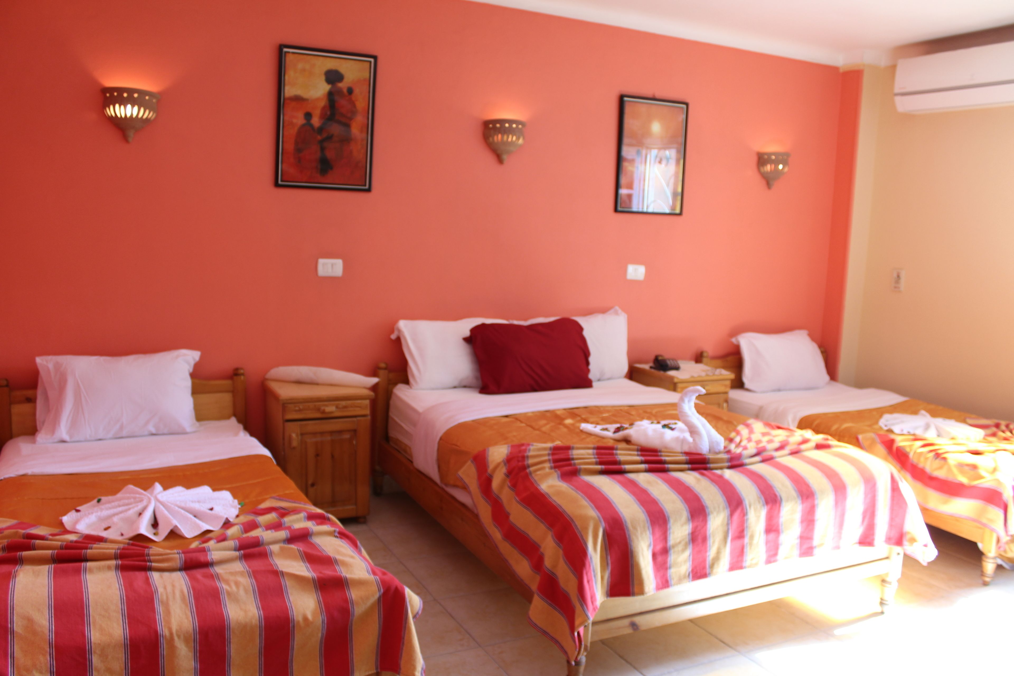 Habitación de hotel con tres camas, cada una con cubrecama a rayas y almohadas blancas. Hay lámparas fijadas a la pared y un cuadro sobre las camas.