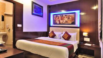 A hotel room with a large bed, a bedside table, a nightstand, a wall-mounted TV, and a framed picture above the bed.