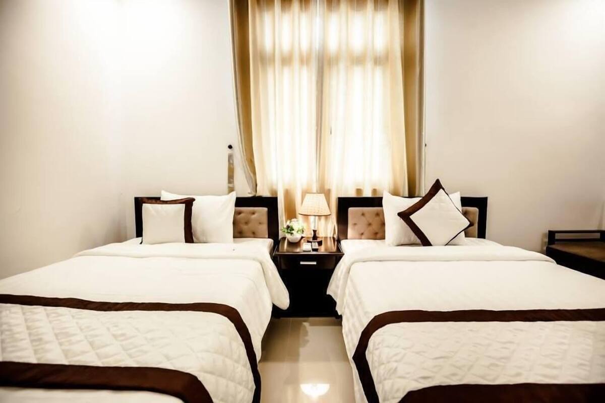 Ein Hotelzimmer mit zwei Betten, einem Nachttisch mit Lampe und Blick auf die Straße durch Vorhänge.
