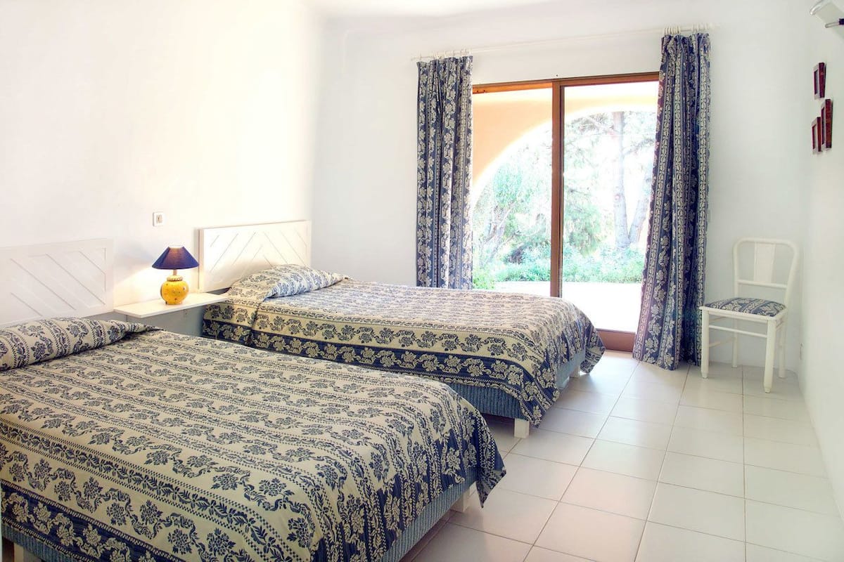 Un dormitorio con dos camas, una mesita de noche, una lámpara, una silla y una ventana con cortinas.