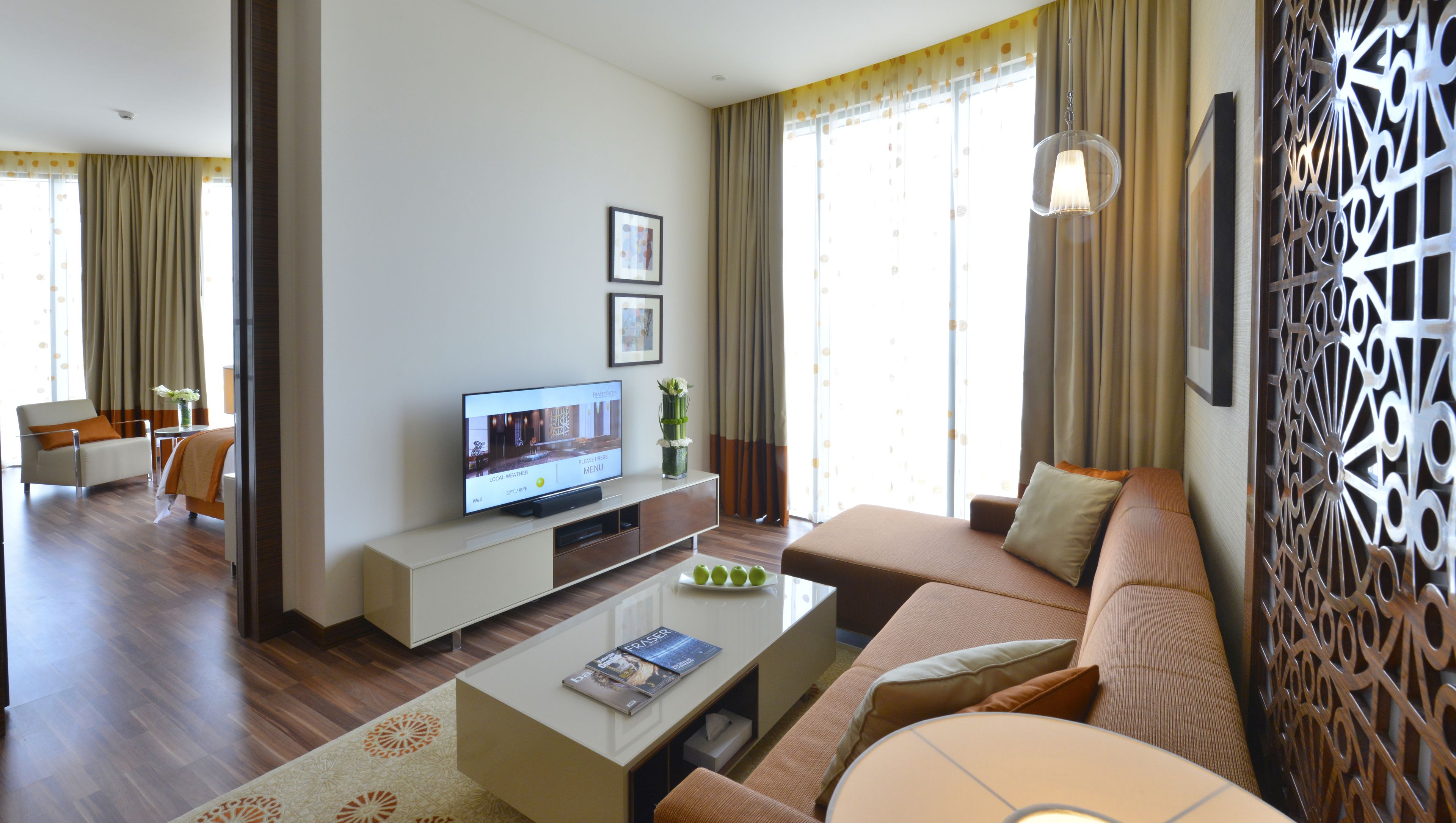 Uma sala de estar moderna com sofá, mesa de centro e televisão. Apresenta um painel decorativo na parede e uma janela com cortinas.