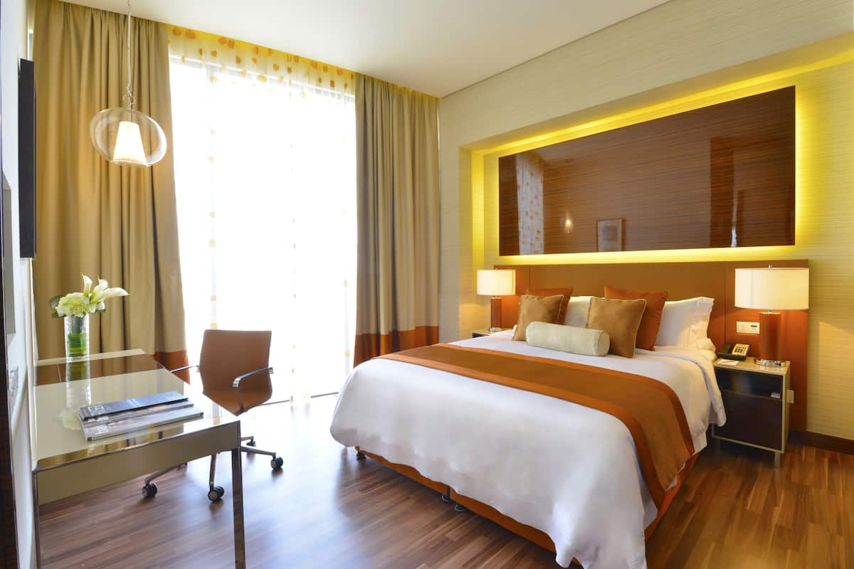 A hotel room with a large bed, bedside lamps, a desk with a chair, and a decorative wall panel.