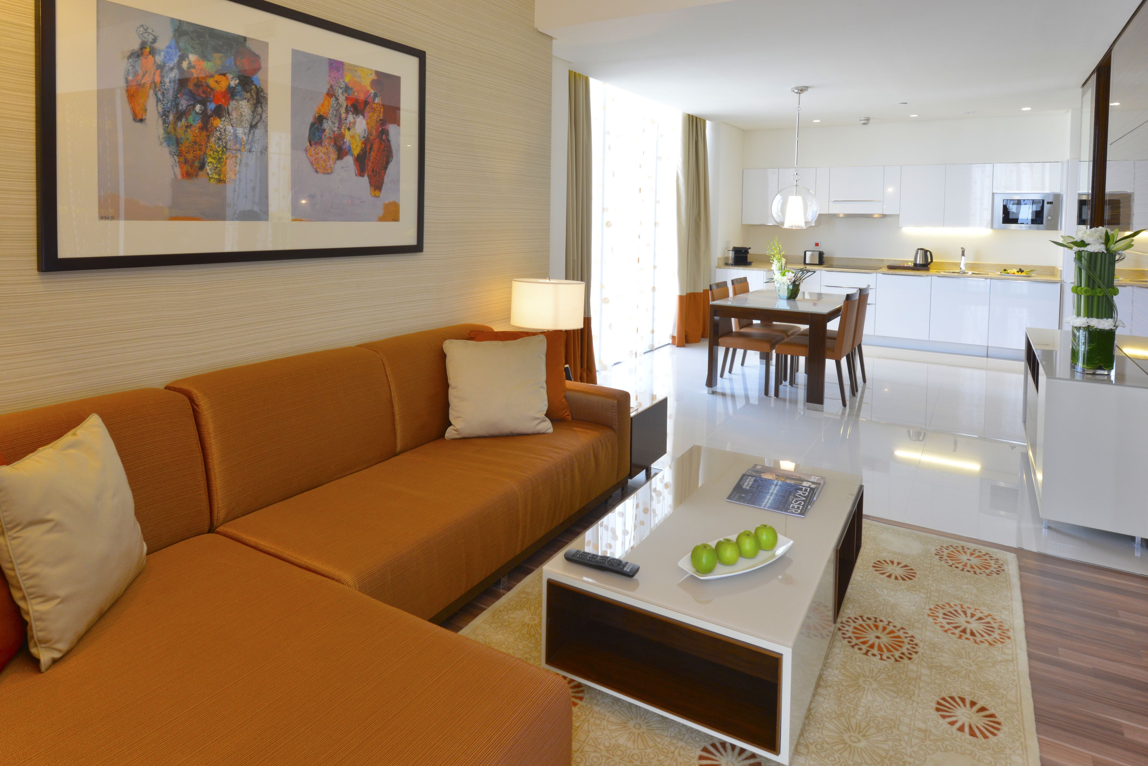 A modern hotel room with a sofa, a coffee table with apples, a framed artwork, and a view into the kitchen and dining area.