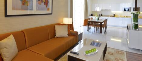 A modern hotel room with a sofa, a coffee table with apples, a framed artwork, and a view into the kitchen and dining area.