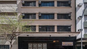 Ein mehrgeschossiges Gebäude mit einer flachen Fassade, Klimaanlagen und einem Eingangsbereich auf Straßenniveau, der mit "Plaza Hotel" beschriftet ist.