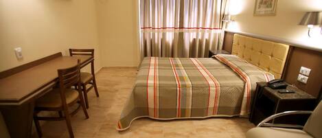 Habitación de hotel con cama, escritorio, silla, televisor y ventana con cortinas.