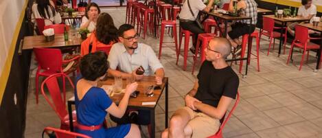 Un restaurante con sillas y mesas rojas, gente cenando, y vista a la calle.