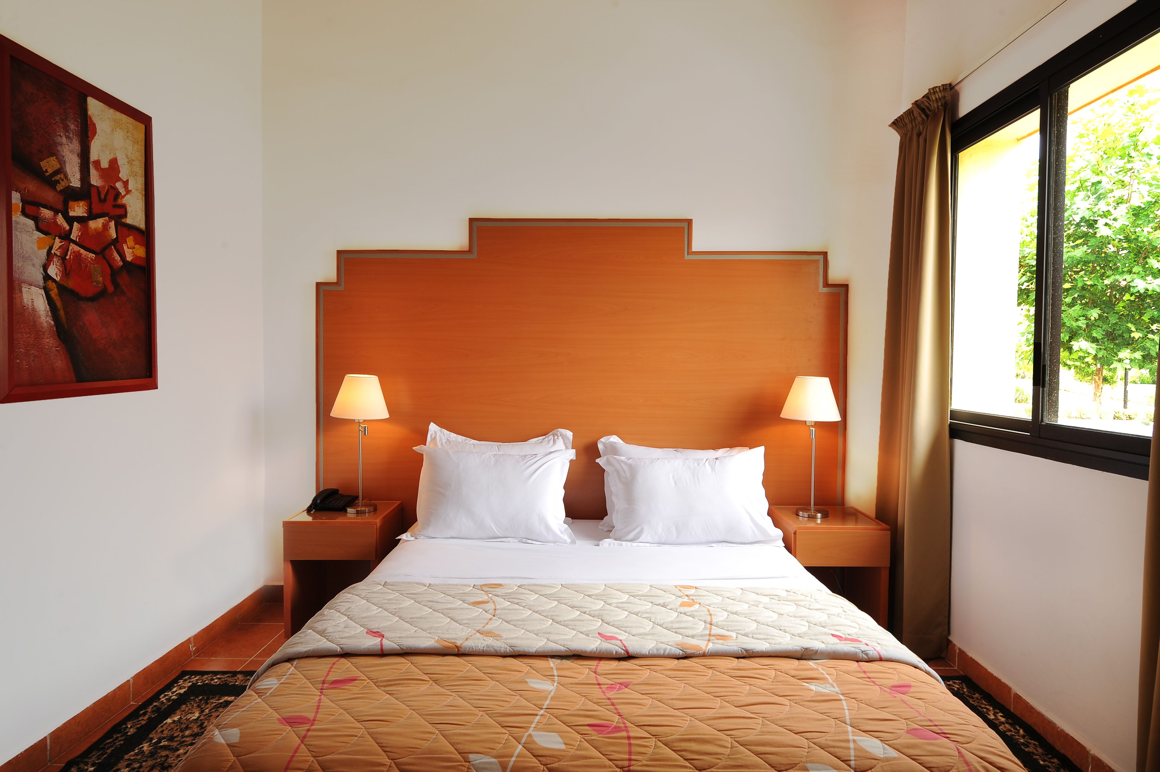 A hotel room with a bed, two bedside lamps, a nightstand, a window with curtains, and a painting on the wall.