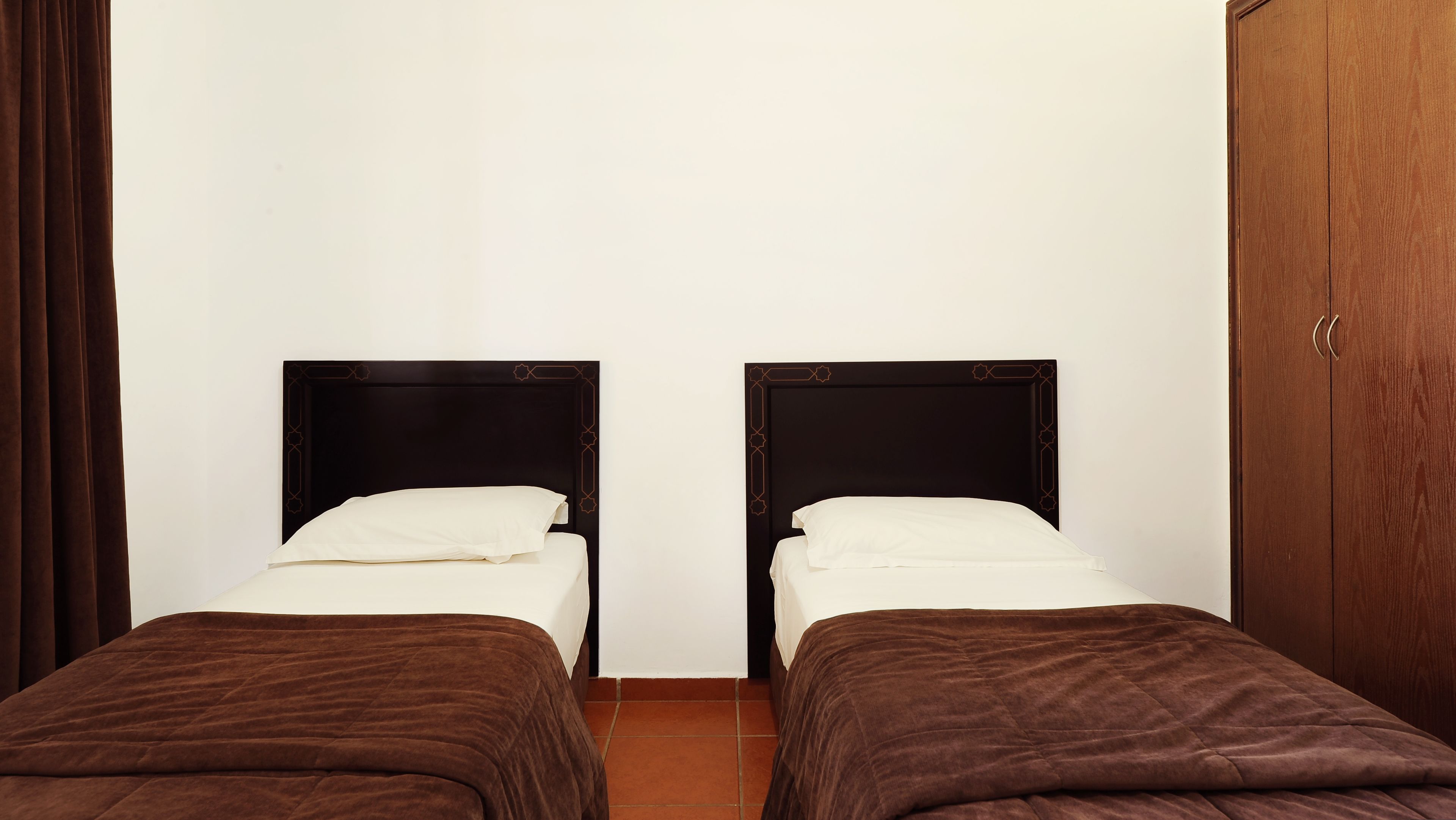 Two single beds with dark wooden headboards and white bedding, in a room with a plain white wall and a wooden door.