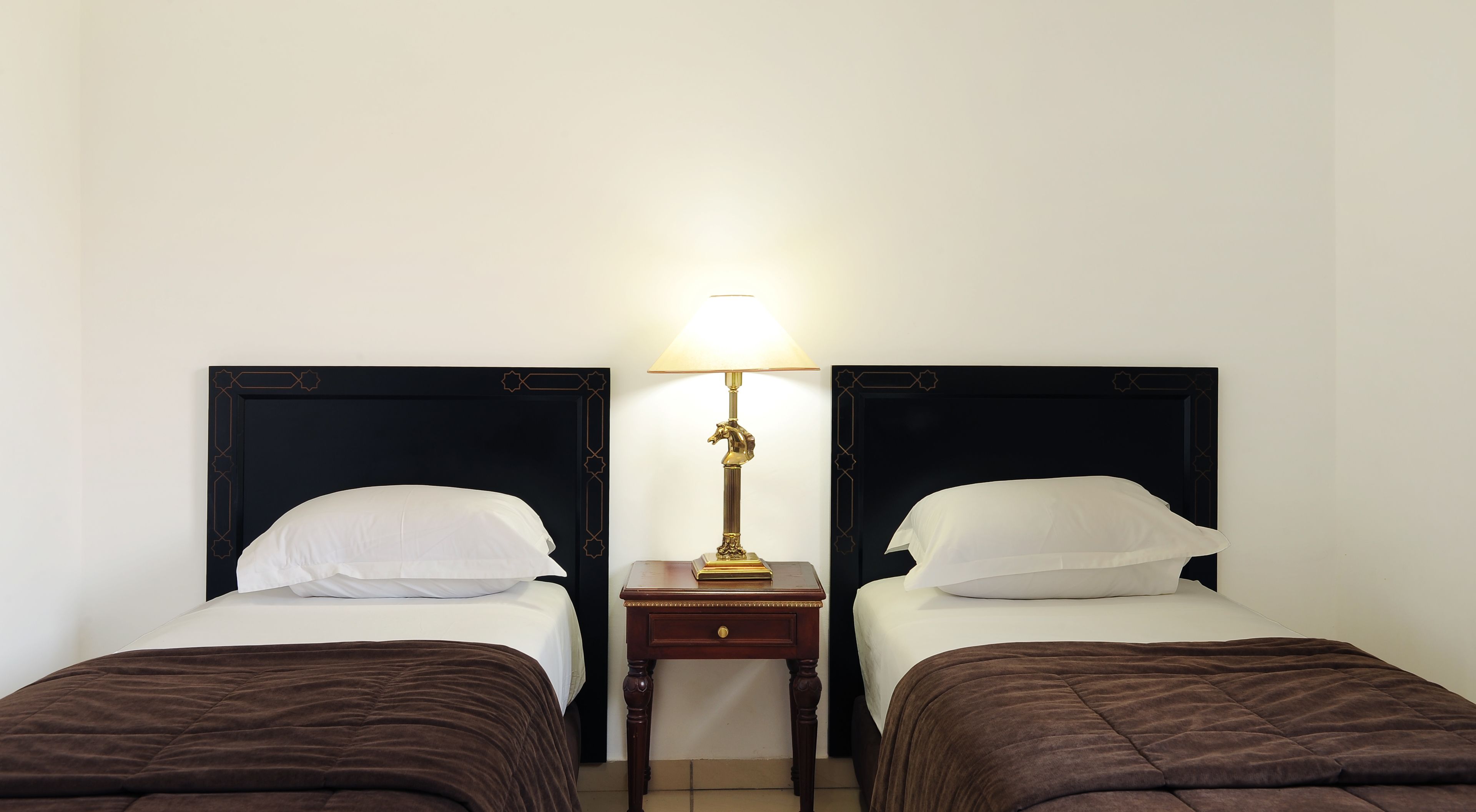 Habitación de hotel con dos camas individuales, cada una con un cabecero oscuro y almohadas blancas. Entre las camas hay una mesita de noche de madera con una lámpara.