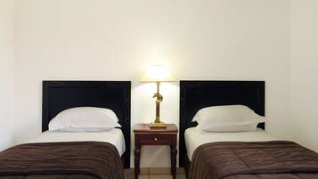 Habitación de hotel con dos camas individuales, cada una con un cabecero oscuro y almohadas blancas. Entre las camas hay una mesita de noche de madera con una lámpara.