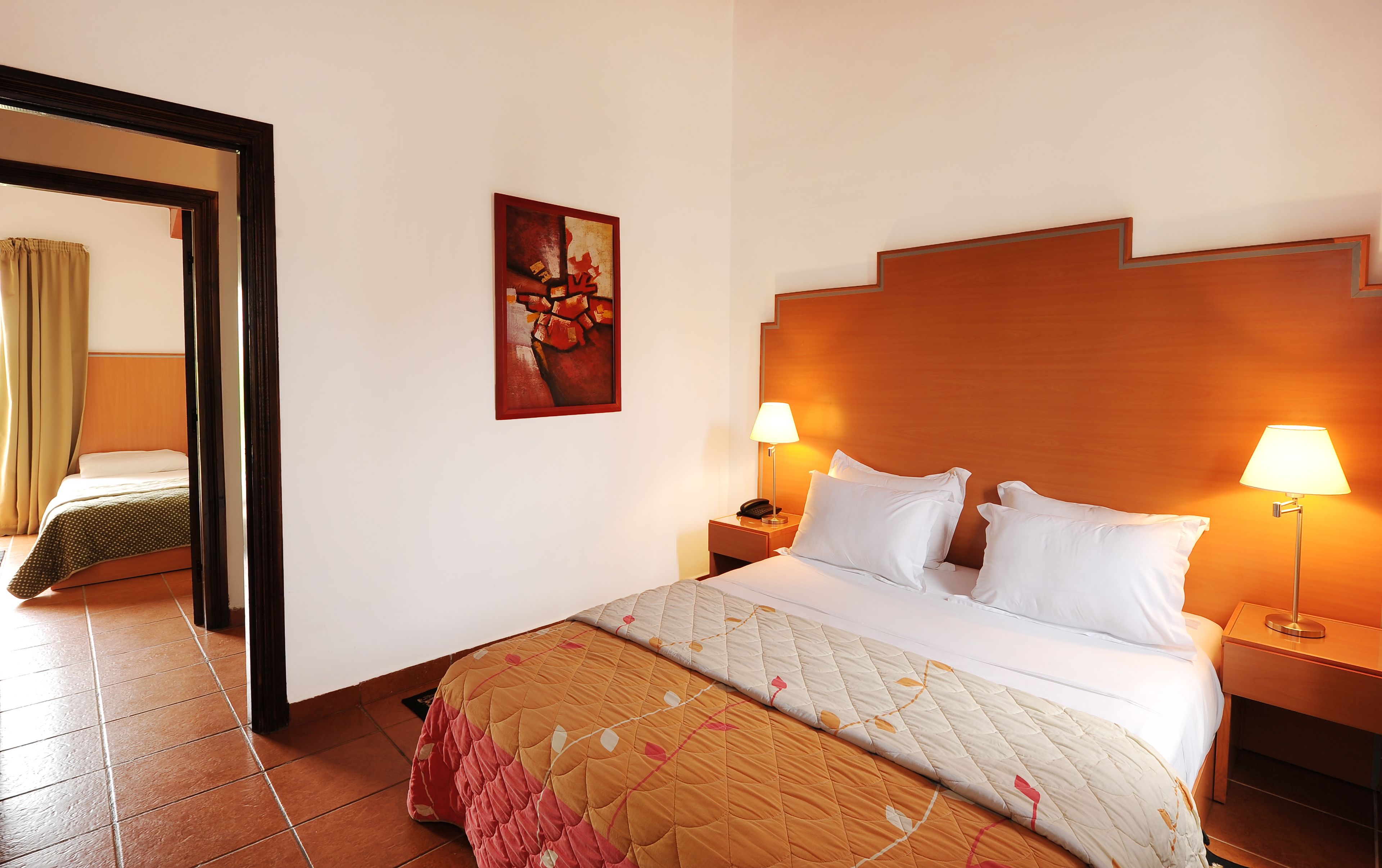 A hotel room with a large bed, two bedside lamps, a nightstand, a painting on the wall, and a mirror reflecting part of the room.