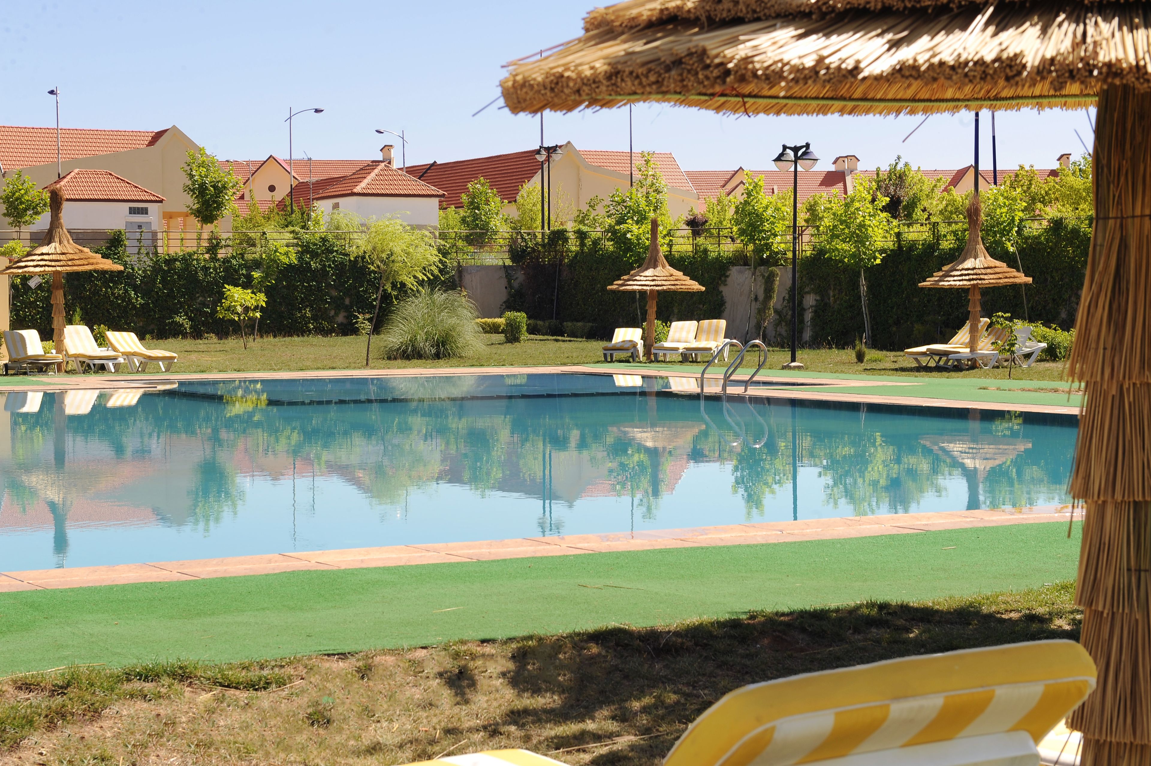 A swimming pool with lounge chairs and umbrellas, surrounded by greenery and buildings with red roofs.