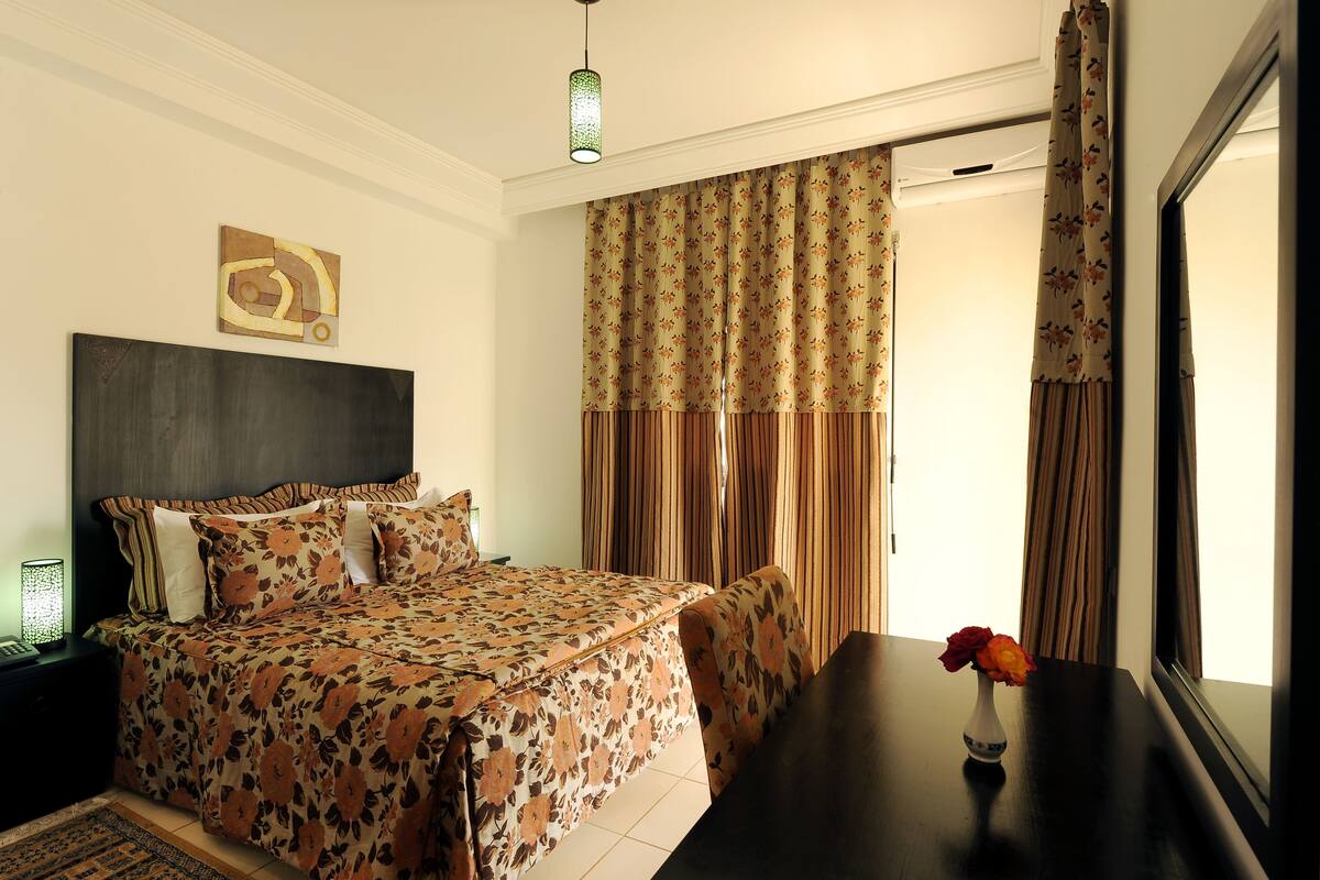 A bedroom with a bed, a bedside table, a vase with flowers, and a window with curtains.