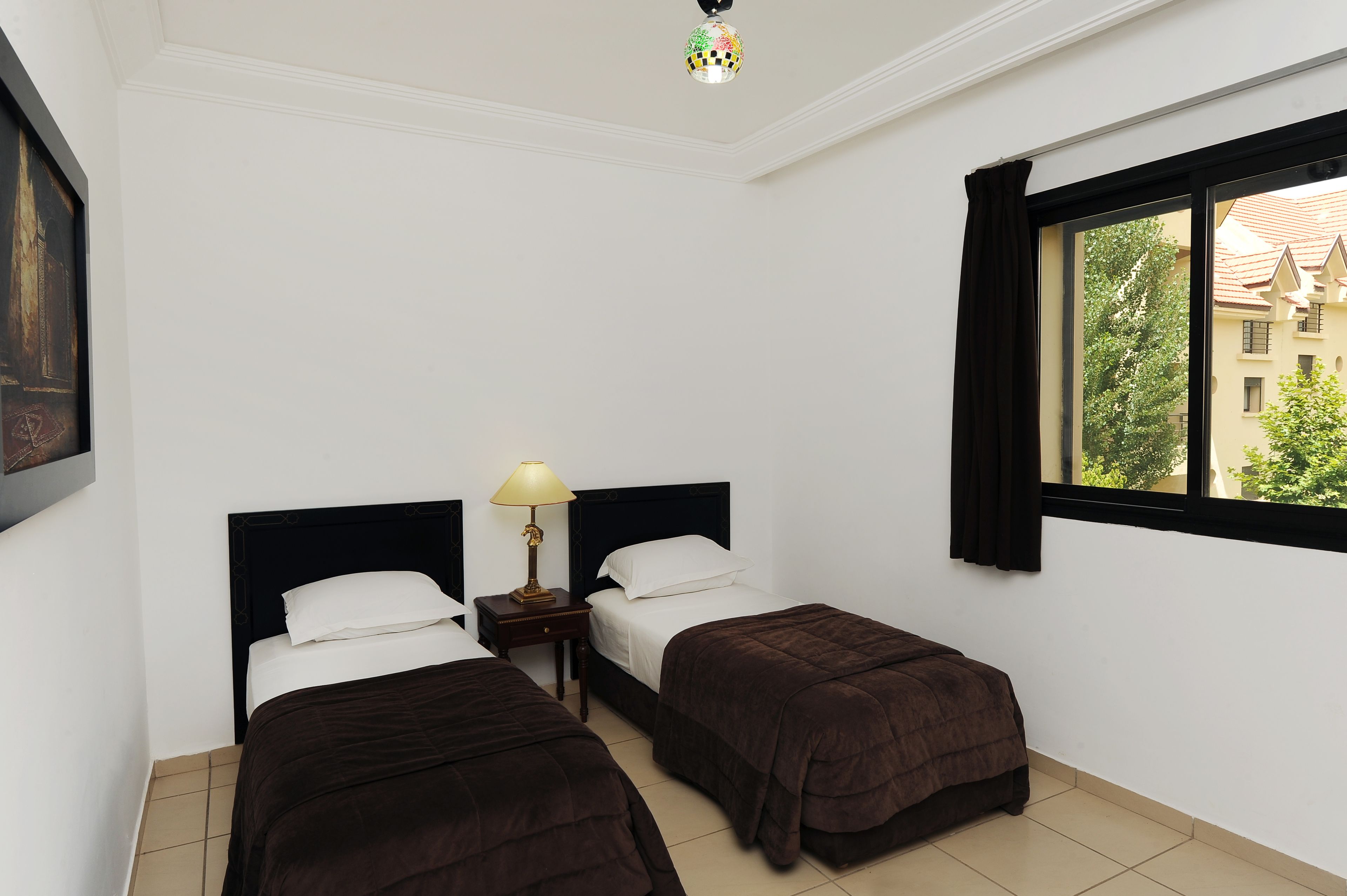 Une chambre d’hôtel avec deux lits, une table de chevet avec une lampe, une fenêtre avec des rideaux et une vue sur les bâtiments environnants.