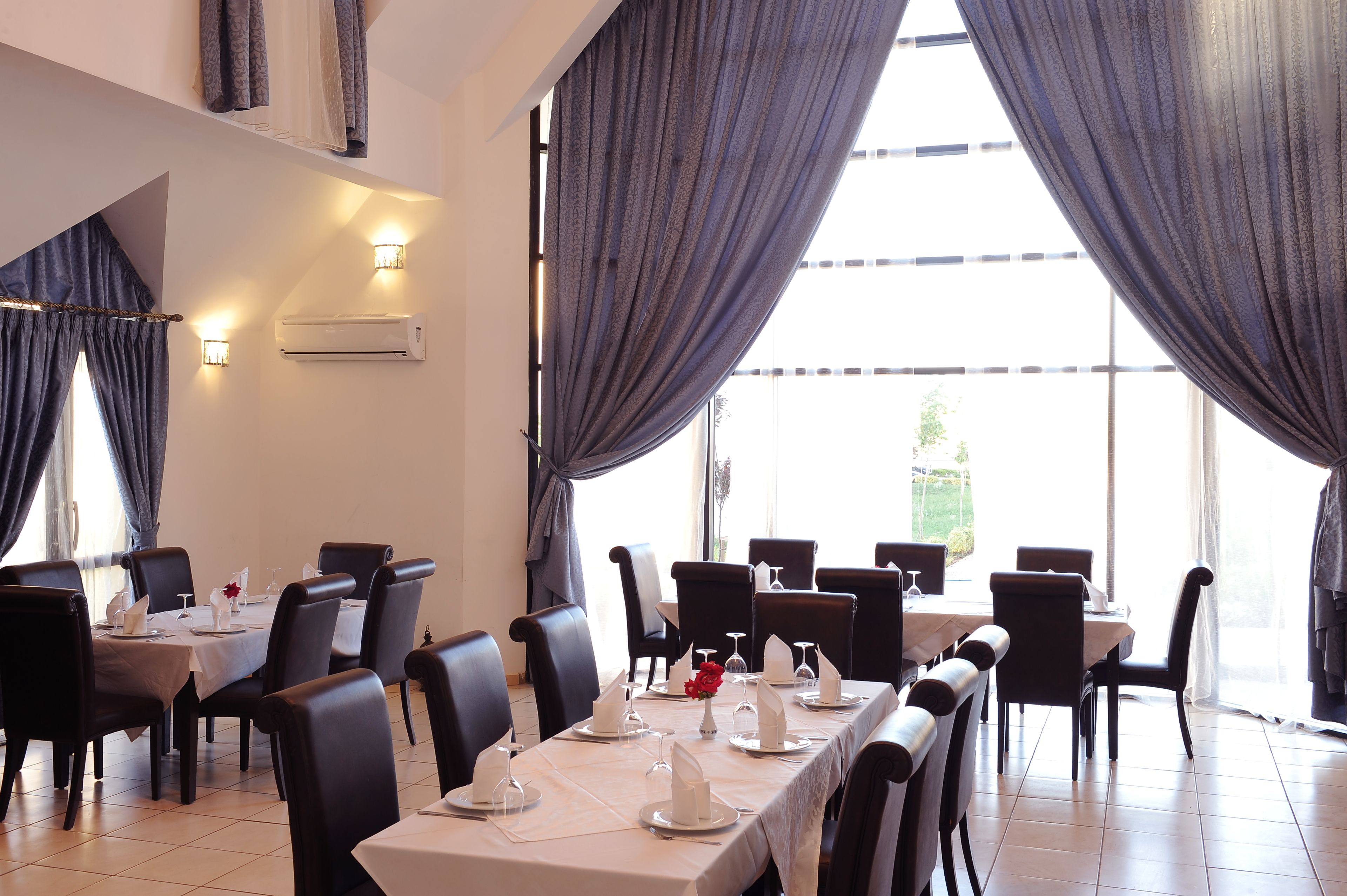A dining area with tables set for four, black chairs, and a large window with curtains.