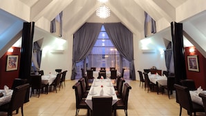 A dining area with a high ceiling, large windows with curtains, and a chandelier. Tables are set with white tablecloths and chairs. - Farah Inn Ifrane (Ifrane)