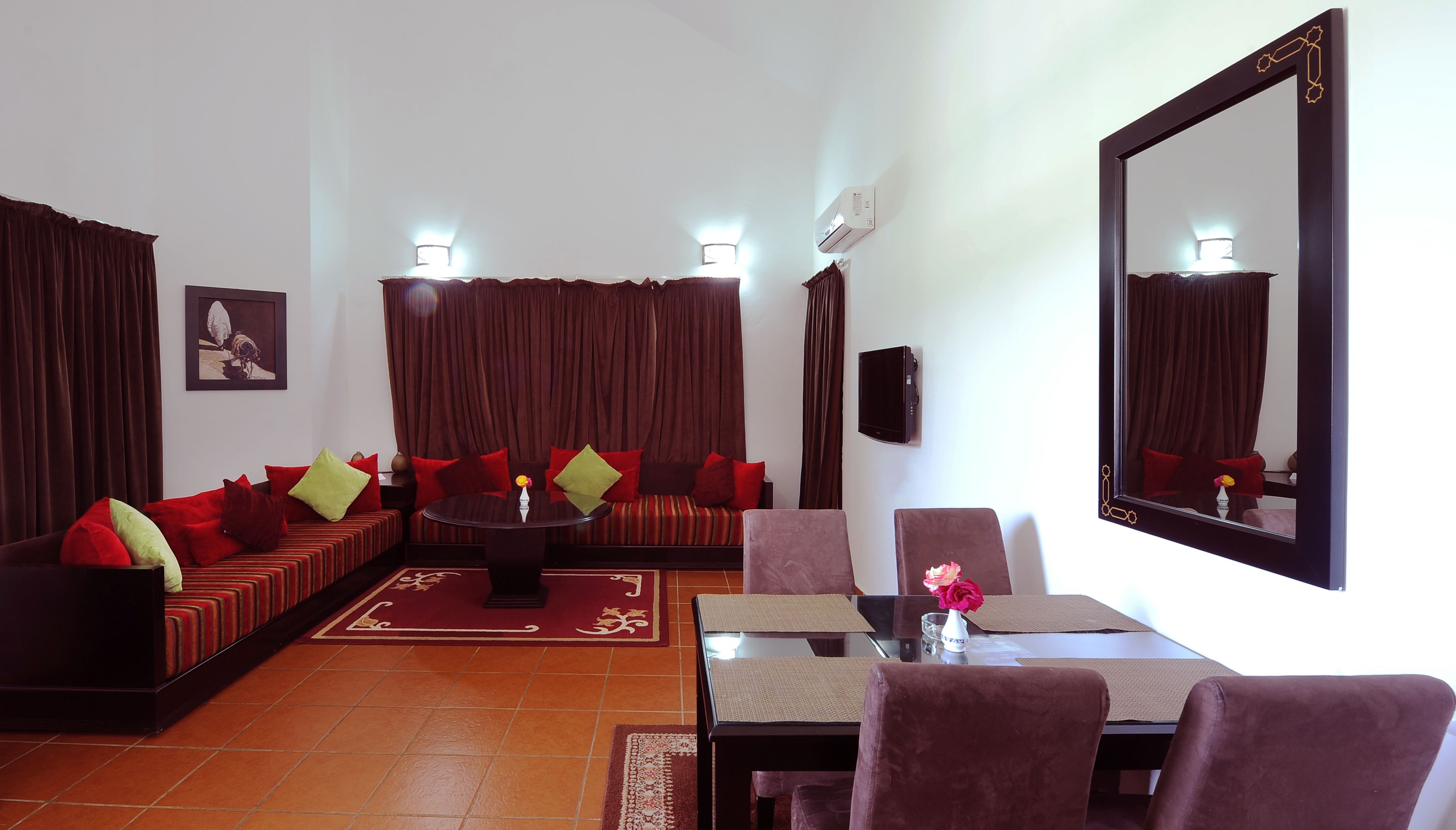 A living room with a sofa set, a dining table with chairs, a large mirror, and a TV on the wall.