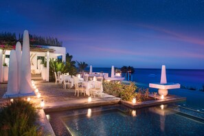 An outdoor dining area with tables and chairs, a pool, and a view of the ocean at night.