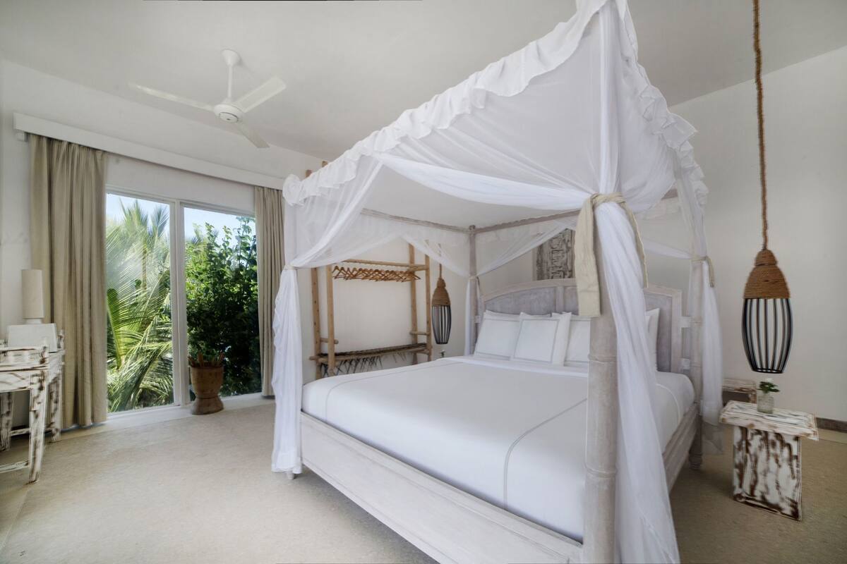 A four-poster canopy bed with a view of palm trees through the window.