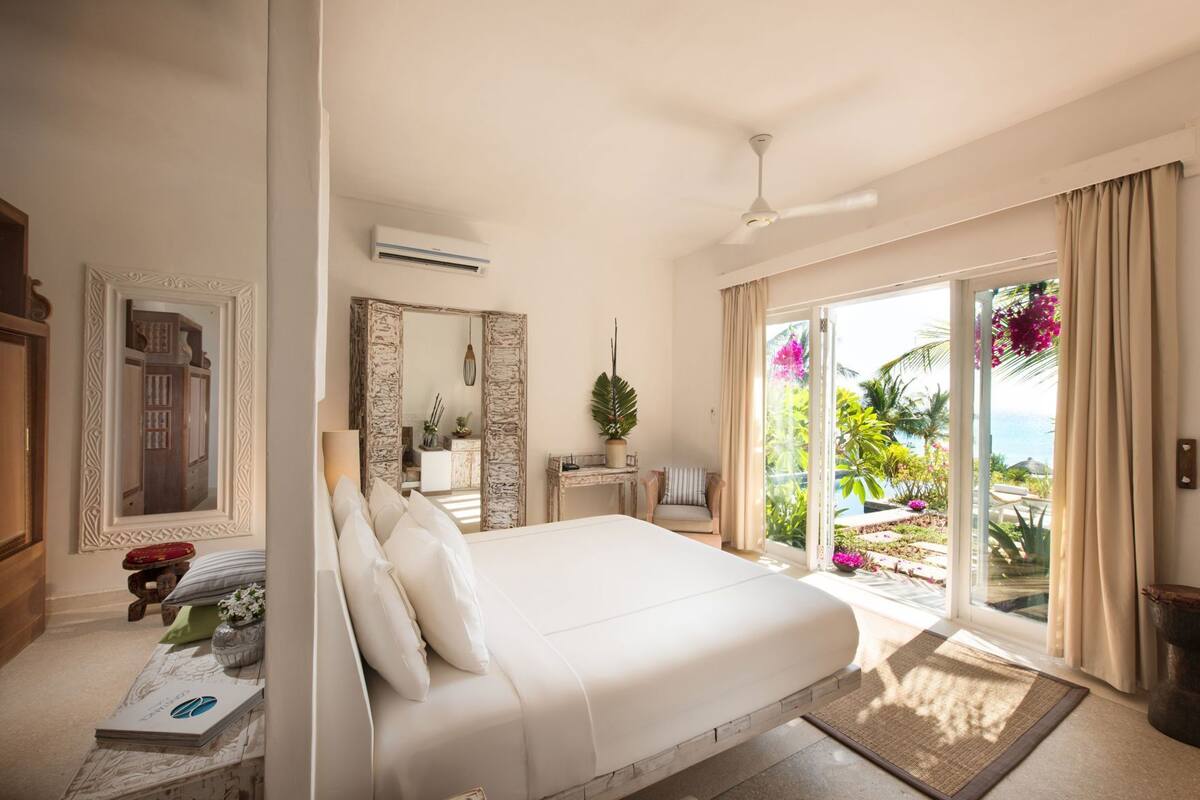 A bedroom with a bed, a mirror, a desk, and a view of a garden through a sliding door.