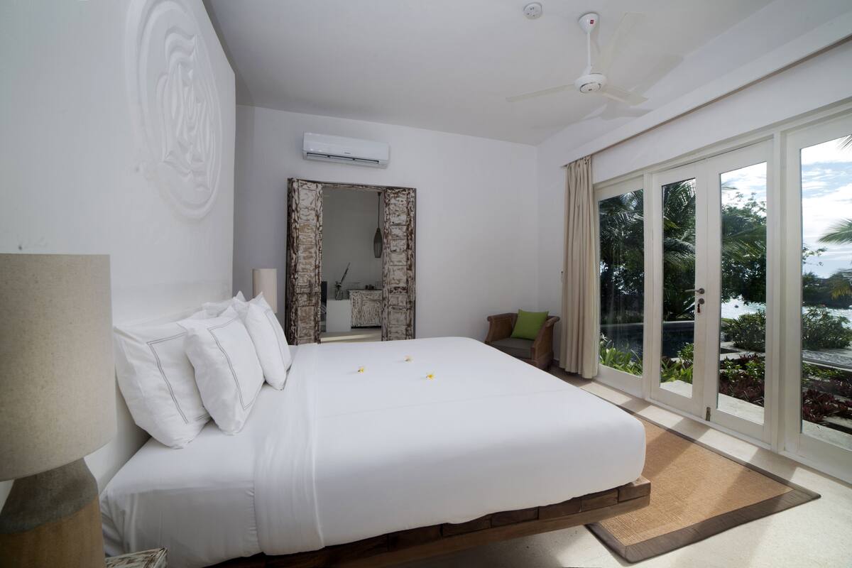 A bedroom with a large bed, a mirror, and a view of the outdoors through large windows.
